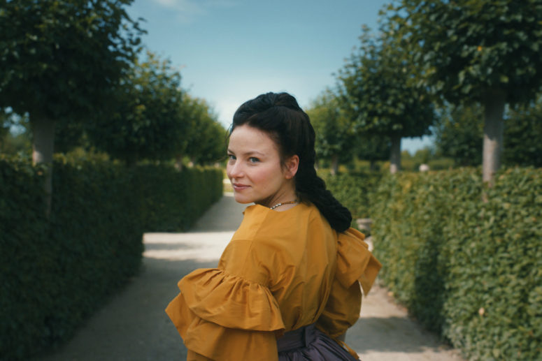 Sisi walks through a park and looks directly over her shoulder at the viewer.